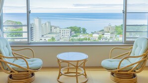 Ein Zimmer mit zwei Korbstühlen und einem kleinen Tisch, ein großes Fenster mit Blick auf Gebäude und das Meer.