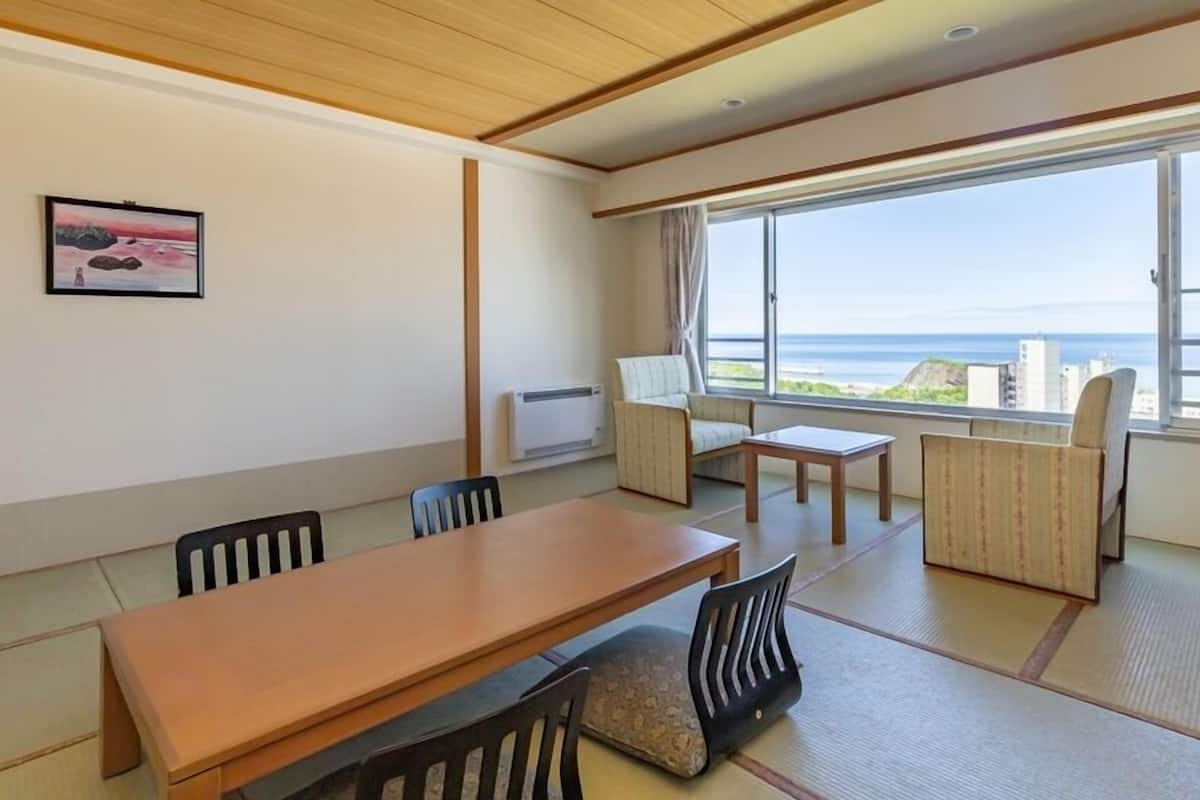 A room with a wooden table, chairs, a small table, a sofa, and a large window with a view of the sea.