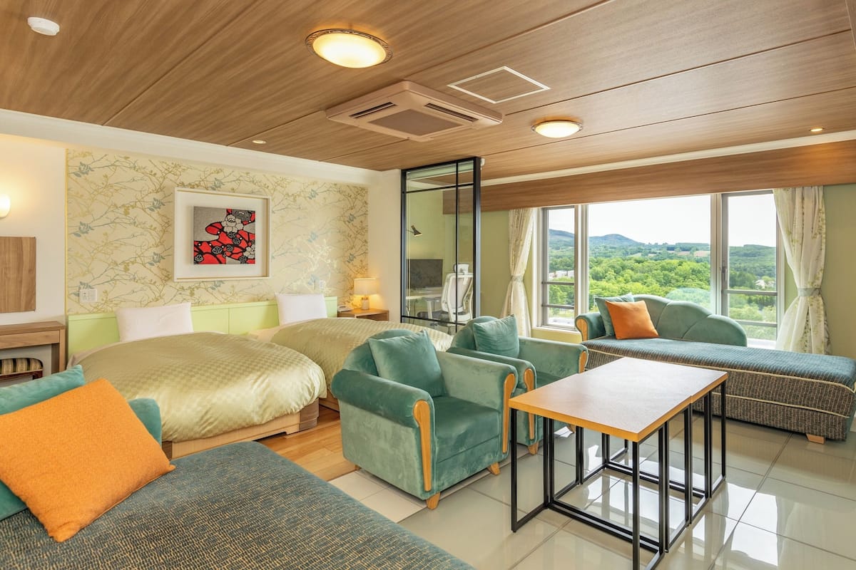 Una habitación de hotel moderna con una cama grande, dos sofás verdes, una mesa de madera y un ventanal con vistas al campo.