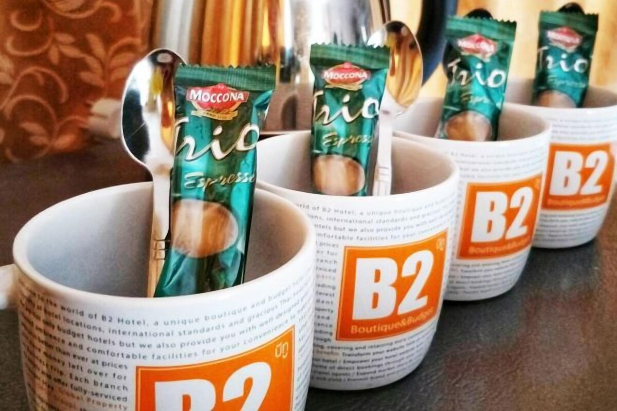 Une rangée de tasses blanches ornées du logo de B2 Boutique&Services, ainsi que des sachets de café expresso Moccona.