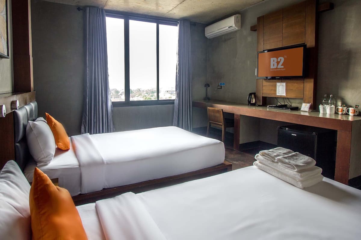 Une chambre d’hôtel avec deux lits, un bureau, une chaise, un appareil de climatisation et une fenêtre avec des rideaux.