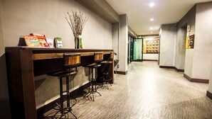 Un área de recepción con mostrador de madera, tres taburetes, un jarrón con ramas y un pasillo que conduce a una habitación con una estantería.