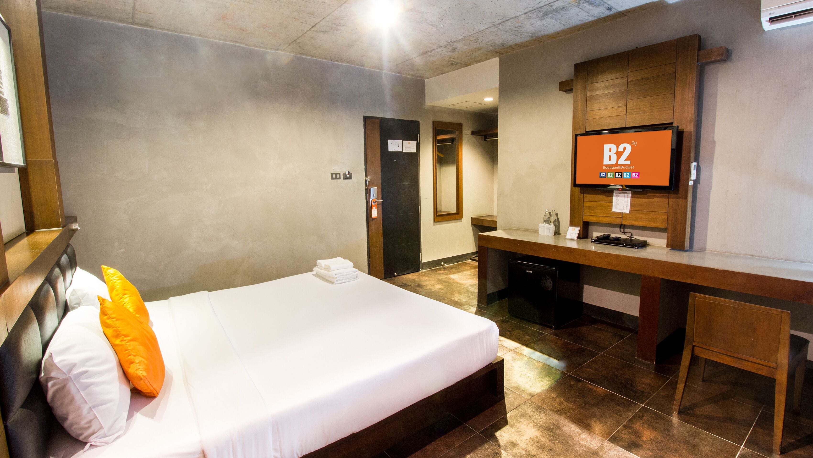 Une chambre d’hôtel équipée d’un lit, d’un bureau, d’un miroir et d’un téléviseur diffusant l’émission « B2 on BoutiqueBudget ».
