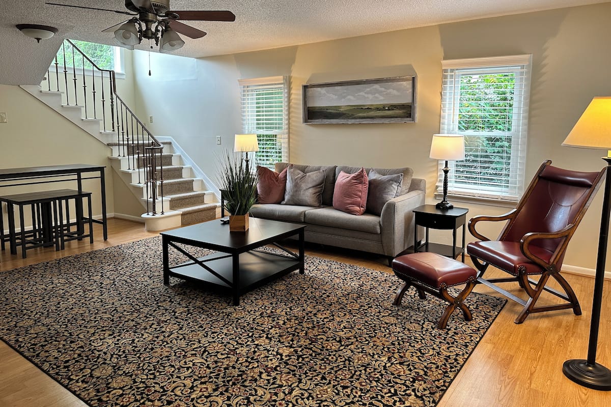 A living room with a sofa, coffee table, and a chair. There is a staircase in the background and a window with blinds.