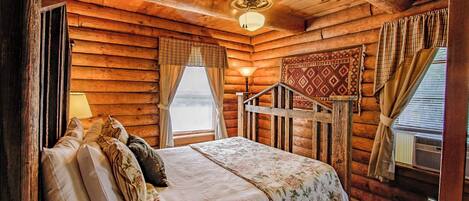 A log cabin bedroom with a bed, pillows, a lamp, a window with curtains, and a ceiling fan.