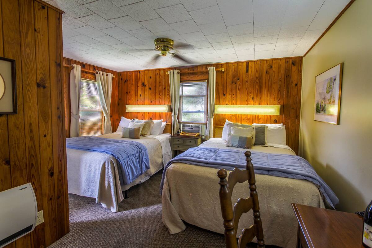 A hotel room with two beds, wooden walls, a ceiling fan, and a painting on the wall.