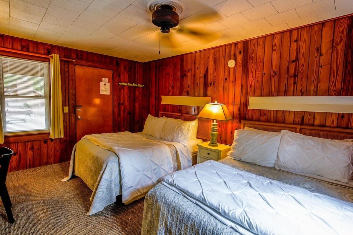 A room with two beds, wooden paneling, a ceiling fan, and a window with curtains.