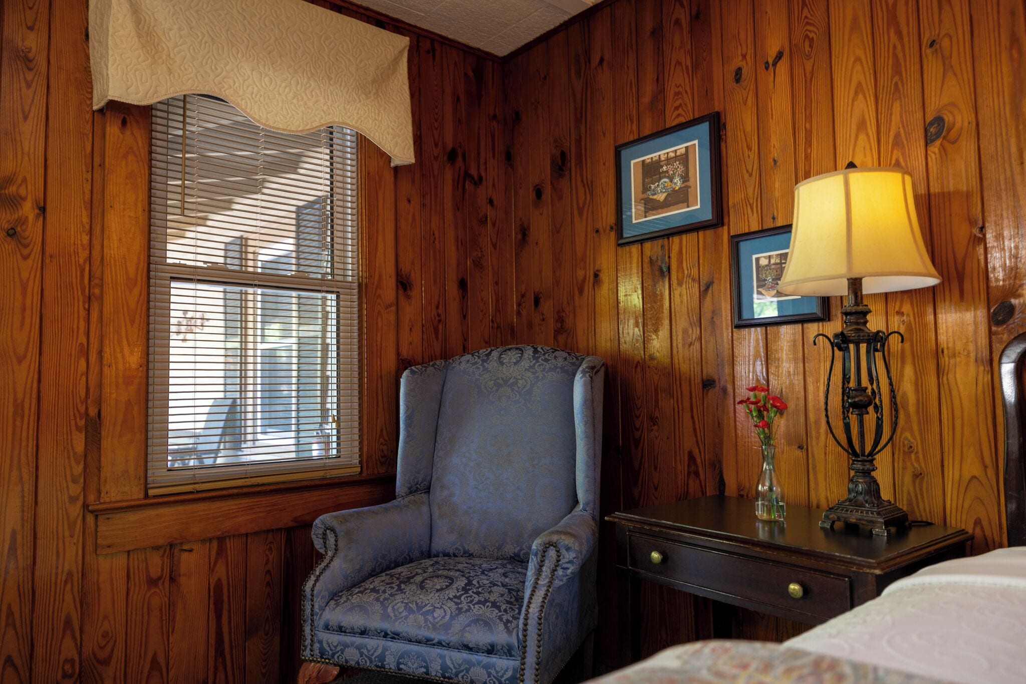 A room with wooden paneled walls, a blue upholstered armchair, a bedside table with a lamp, and a framed picture on the wall.