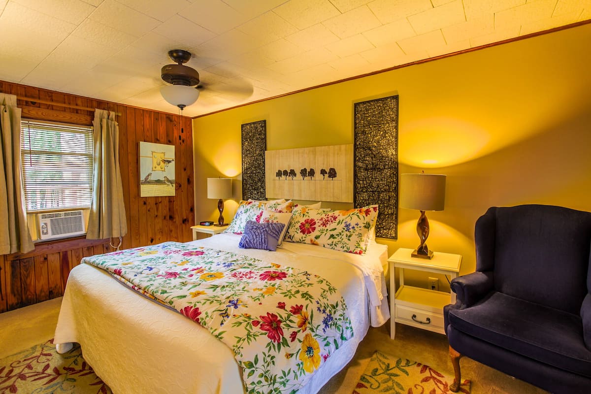 A bedroom with a large bed, two bedside tables, a chair, a ceiling fan, and a window with curtains.