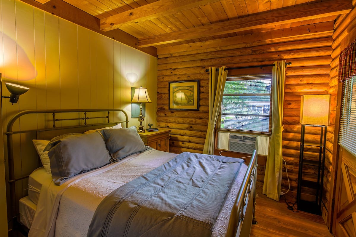 A wooden cabin bedroom with a bed, bedside lamps, a framed picture, and a window with curtains.