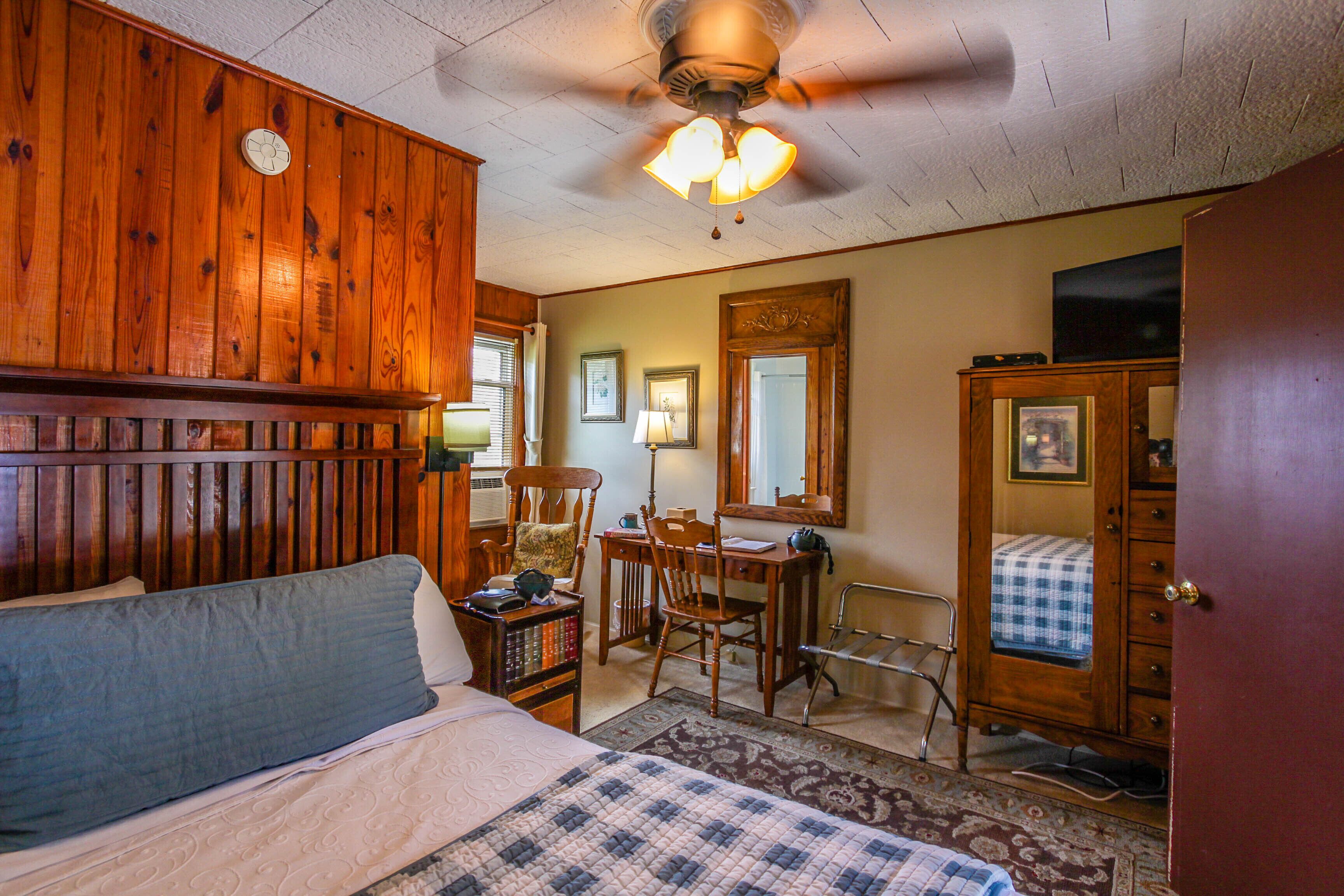 A room with a bed, wooden paneling, a ceiling fan, a desk with a chair, a mirror, and a wardrobe.