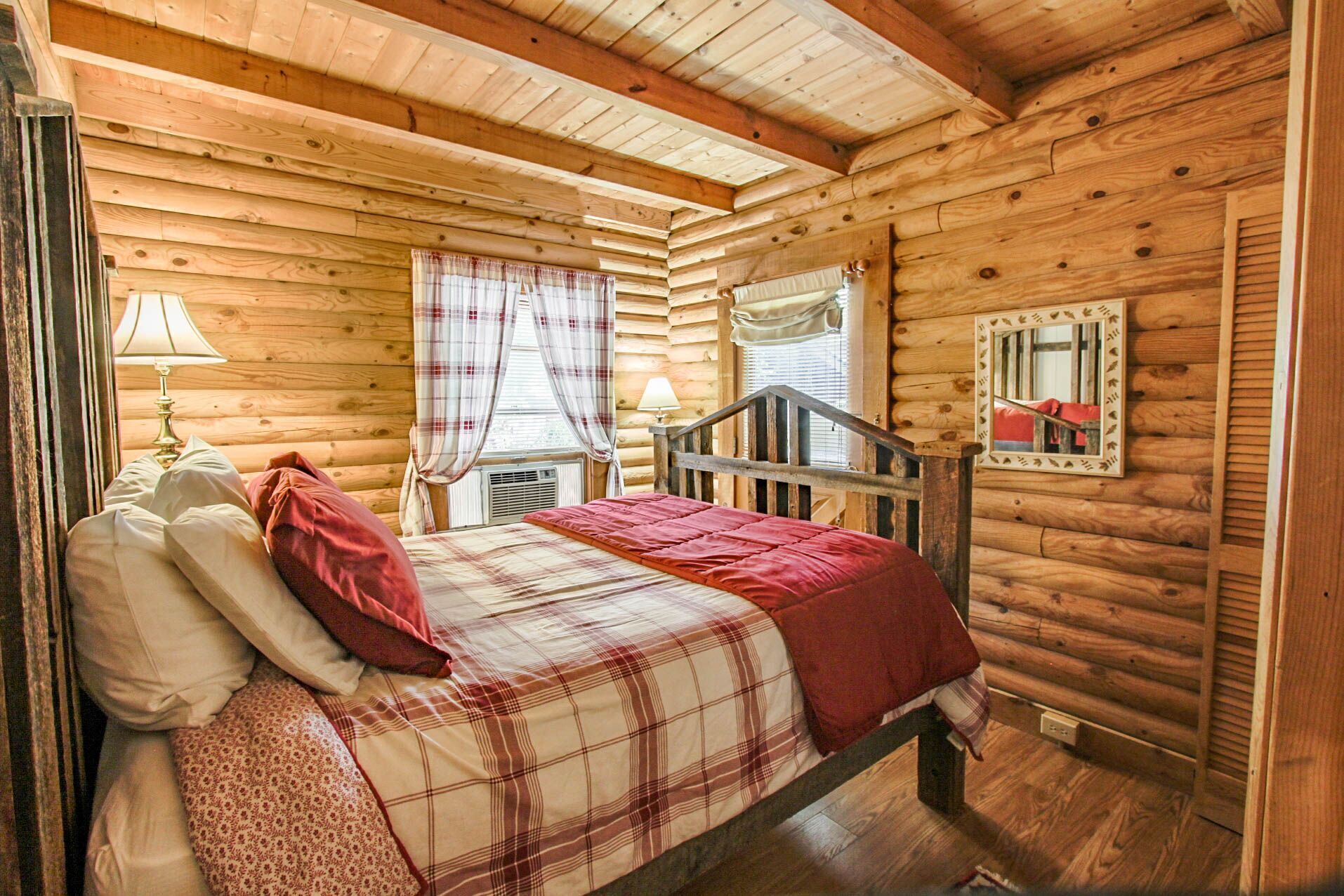 Une chambre confortable dans un chalet en rondins, avec un lit, une table de chevet, une lampe, une fenêtre avec des rideaux et un miroir au mur.