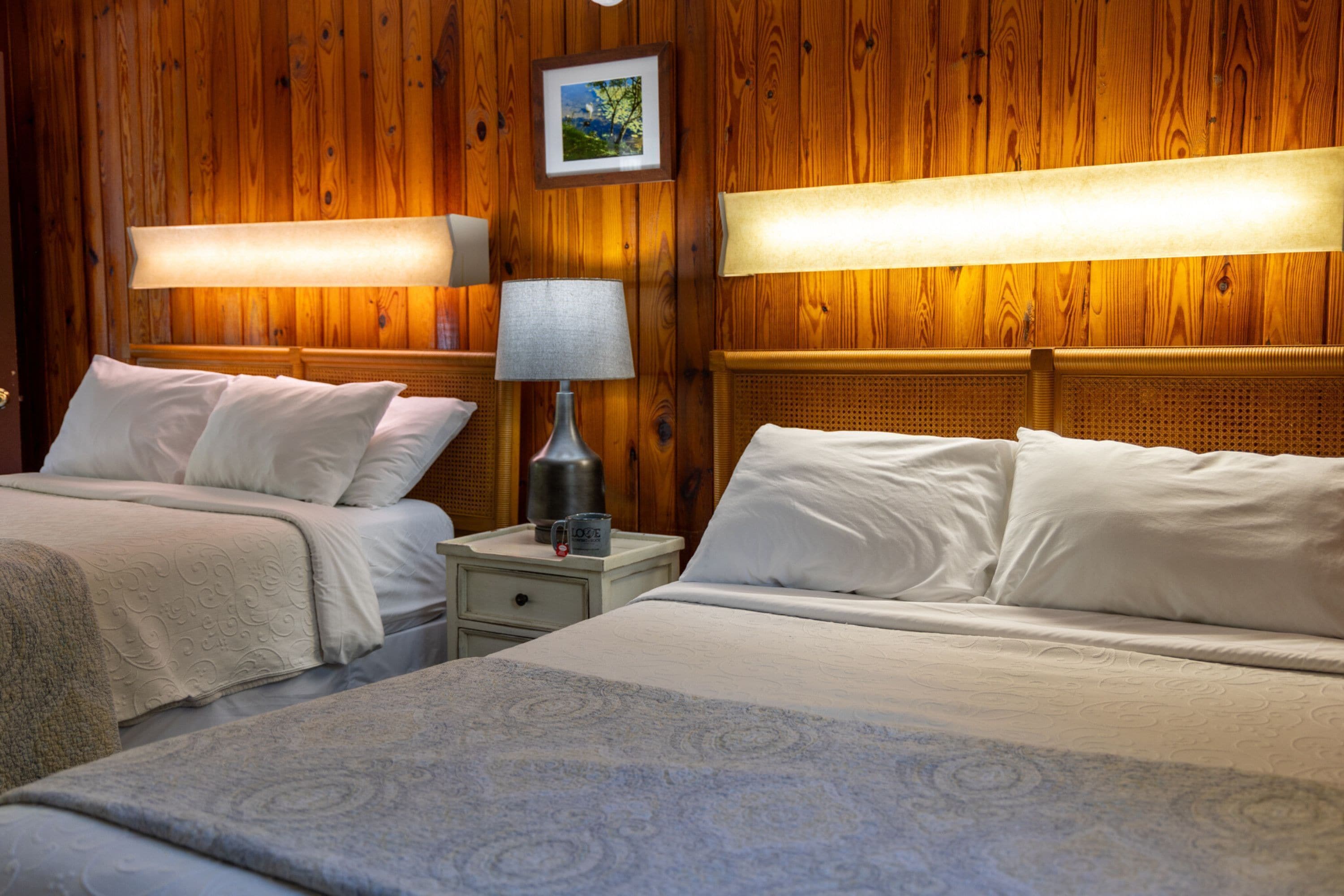 Hotellihuone, jossa on kaksi sänkyä, puupanelointi, seinällä oleva kehystetty taulu ja yöpöytävalaisimet.