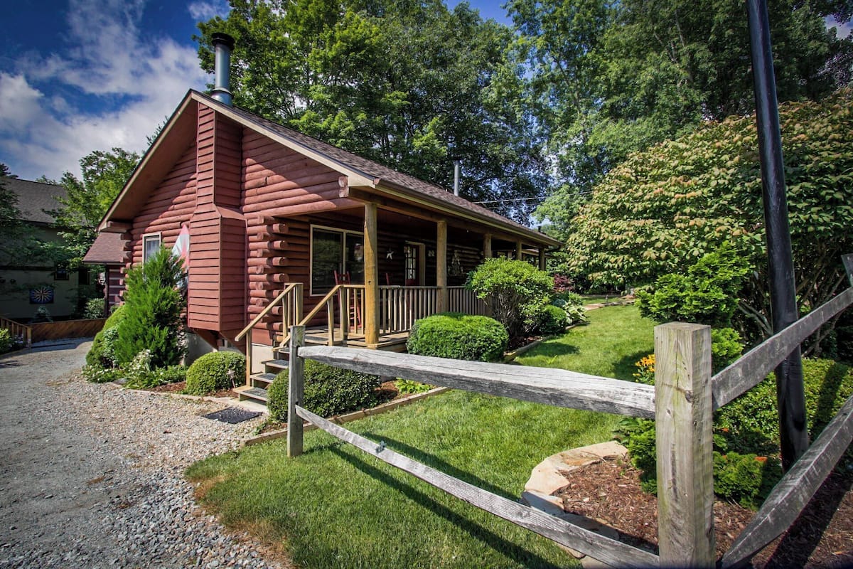 A log cabin with a porch, surrounded by a wooden fence and lush greenery.