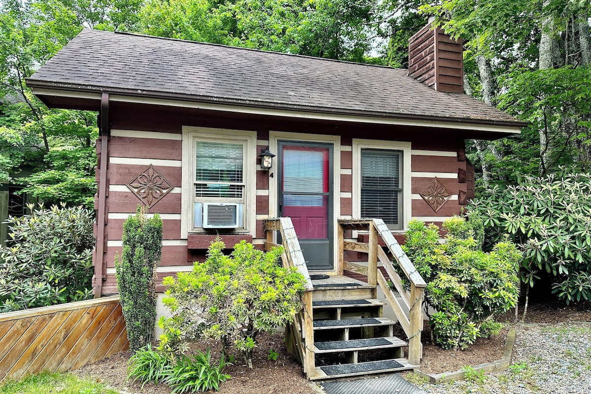 A small, single-story cabin with a red door, two windows with shutters, and a small porch with steps leading up to the door.