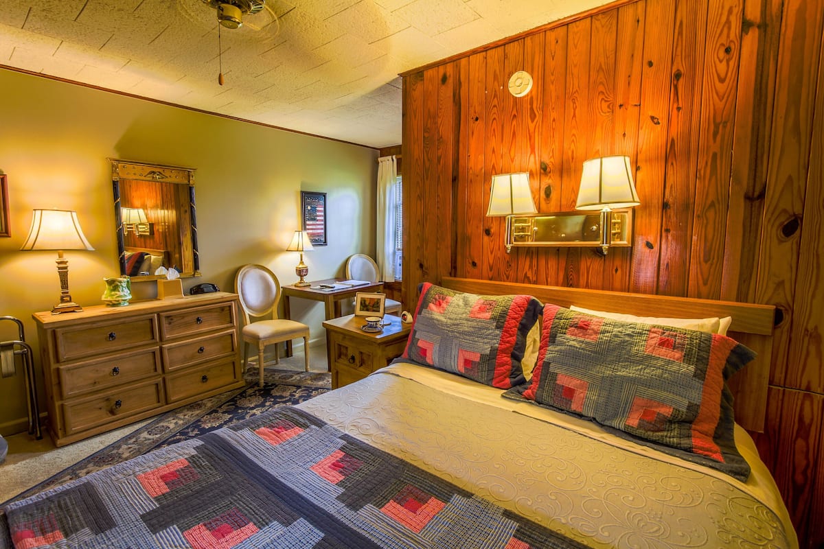 A bedroom with a wooden headboard, a bed with a quilt, a desk with a chair, a dresser, and two wall-mounted lamps.
