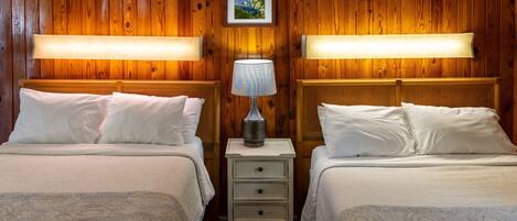 A room with two beds, each with a headboard, a bedside table with a lamp, and a framed picture on the wall.