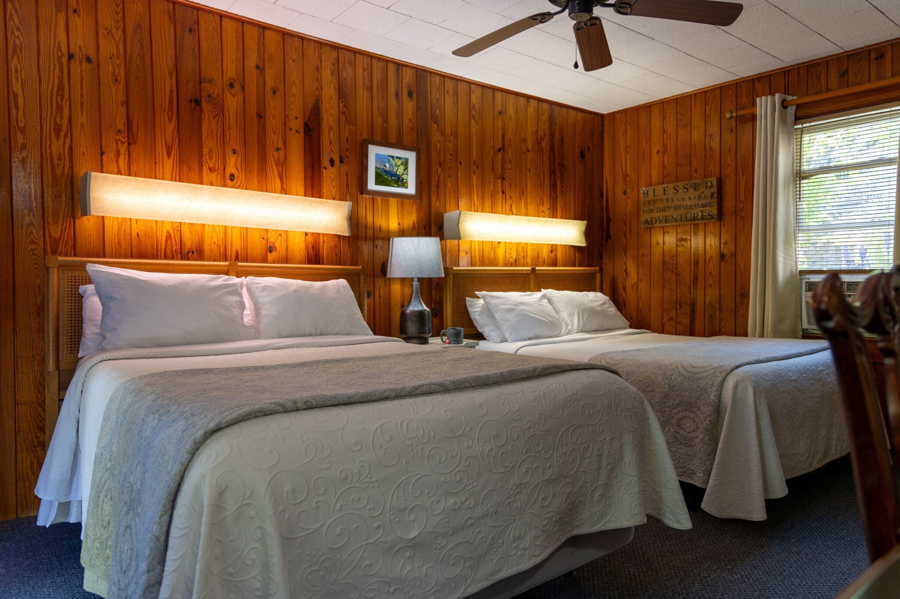A room with two beds, wooden walls, ceiling fan, and a window with curtains.