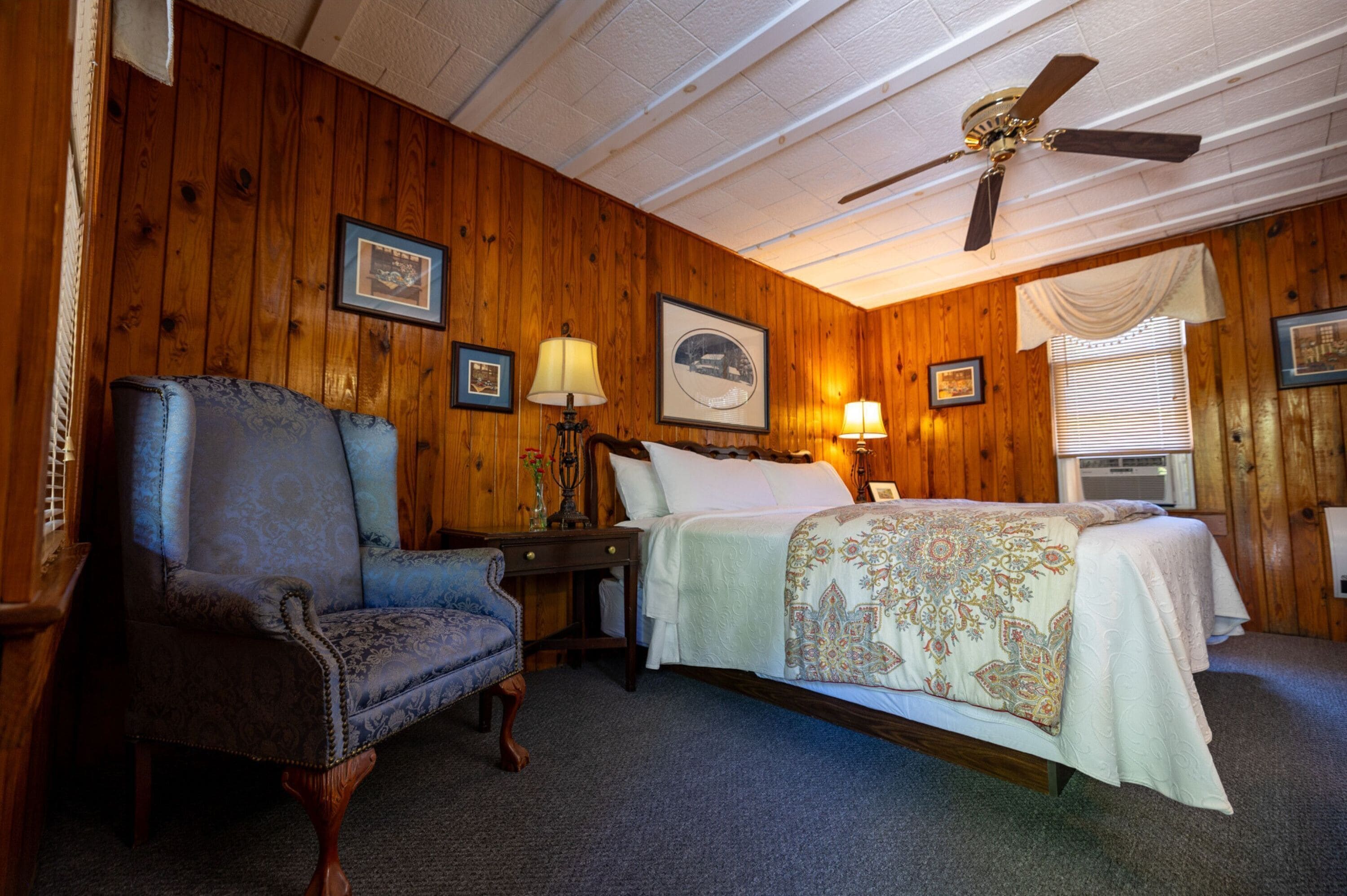 A wooden-paneled room with a bed, a chair, a nightstand, and a ceiling fan.