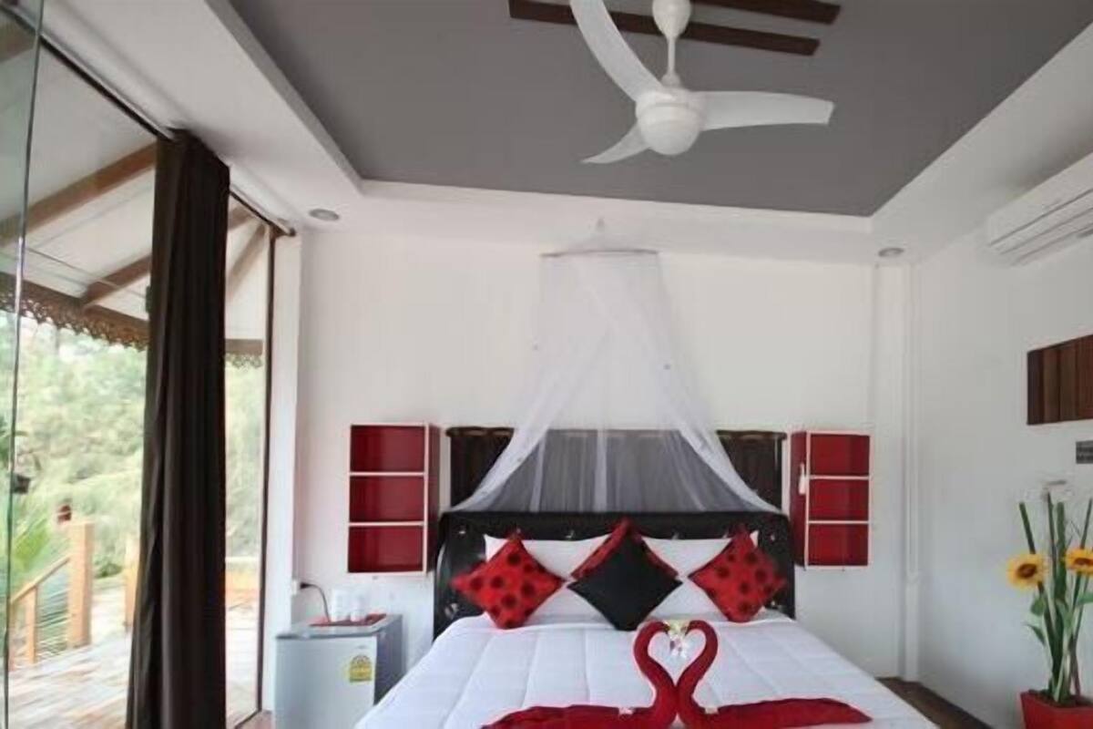 A bedroom with a bed, a ceiling fan, a mosquito net, a wall-mounted air conditioner, and a window with curtains.