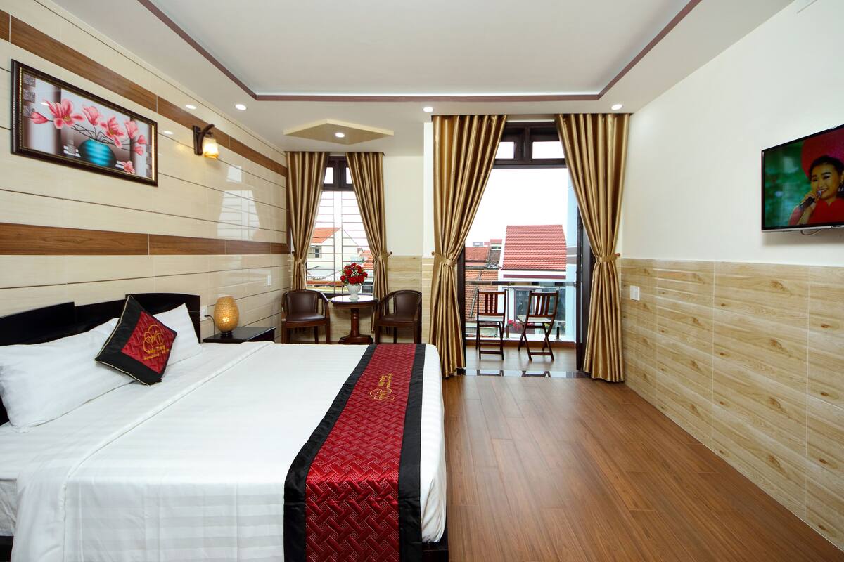 Habitación de hotel con una cama grande, televisión, balcón con mesa y sillas, y vista a la ciudad.