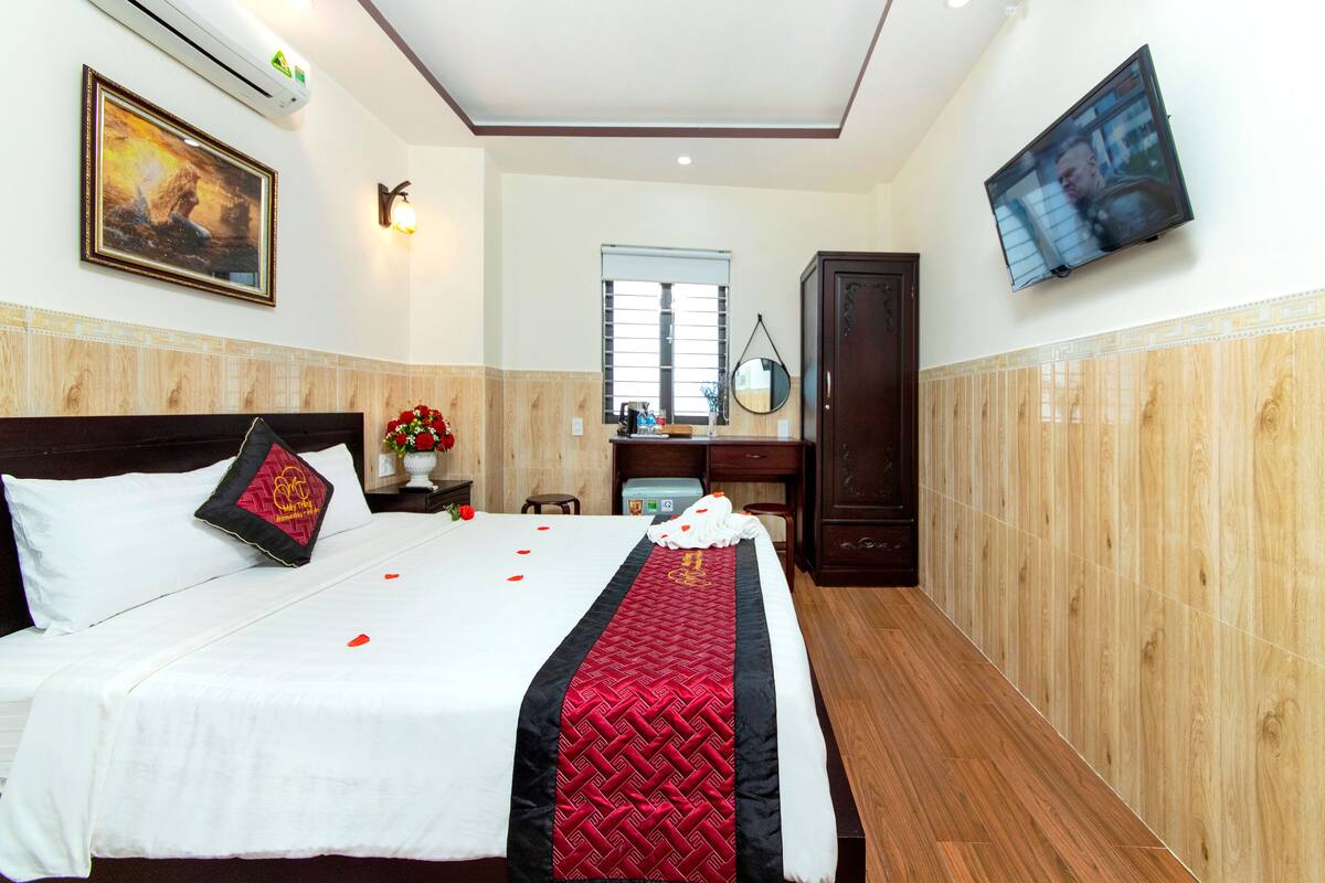 Una cama bien hecha con colcha roja y negra, un armario de madera, un televisor montado en la pared y una ventana con persianas.