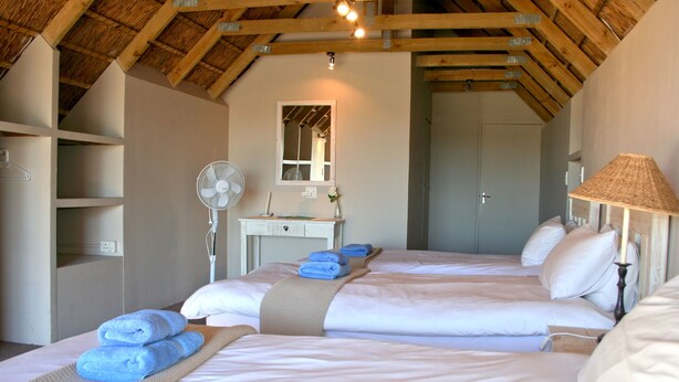 Quarto com duas camas, uma escrivaninha, um espelho, um ventilador e um abajur.