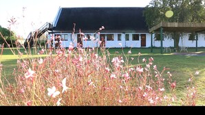 Um edifício com fachada branca e telhado preto, rodeado por um espaço gramado com flores cor-de-rosa.