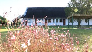 Um edifício branco com telhado escuro, uma escadaria e uma área gramada com flores cor-de-rosa na frente.