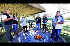 A group of people gathered around a fire pit in an outdoor setting with a covered area and chairs in the background. - Zoutpan Struishuis (Albertinia)