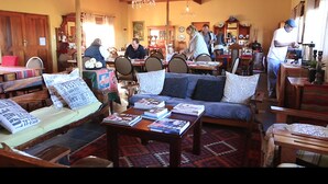 Uma sala de estar aconchegante com um sofá, cadeiras e uma mesa de centro com livros. Ao fundo, há uma área de jantar com pessoas sentadas em mesas.