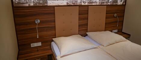 A hotel room with a wooden headboard, two beds with white linens, a wall-mounted picture of a town, and a power outlet on the nightstand.
