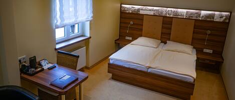 A bedroom with a wooden bed, a desk with a chair, a window with sheer curtains, and a wall-mounted nightstand.