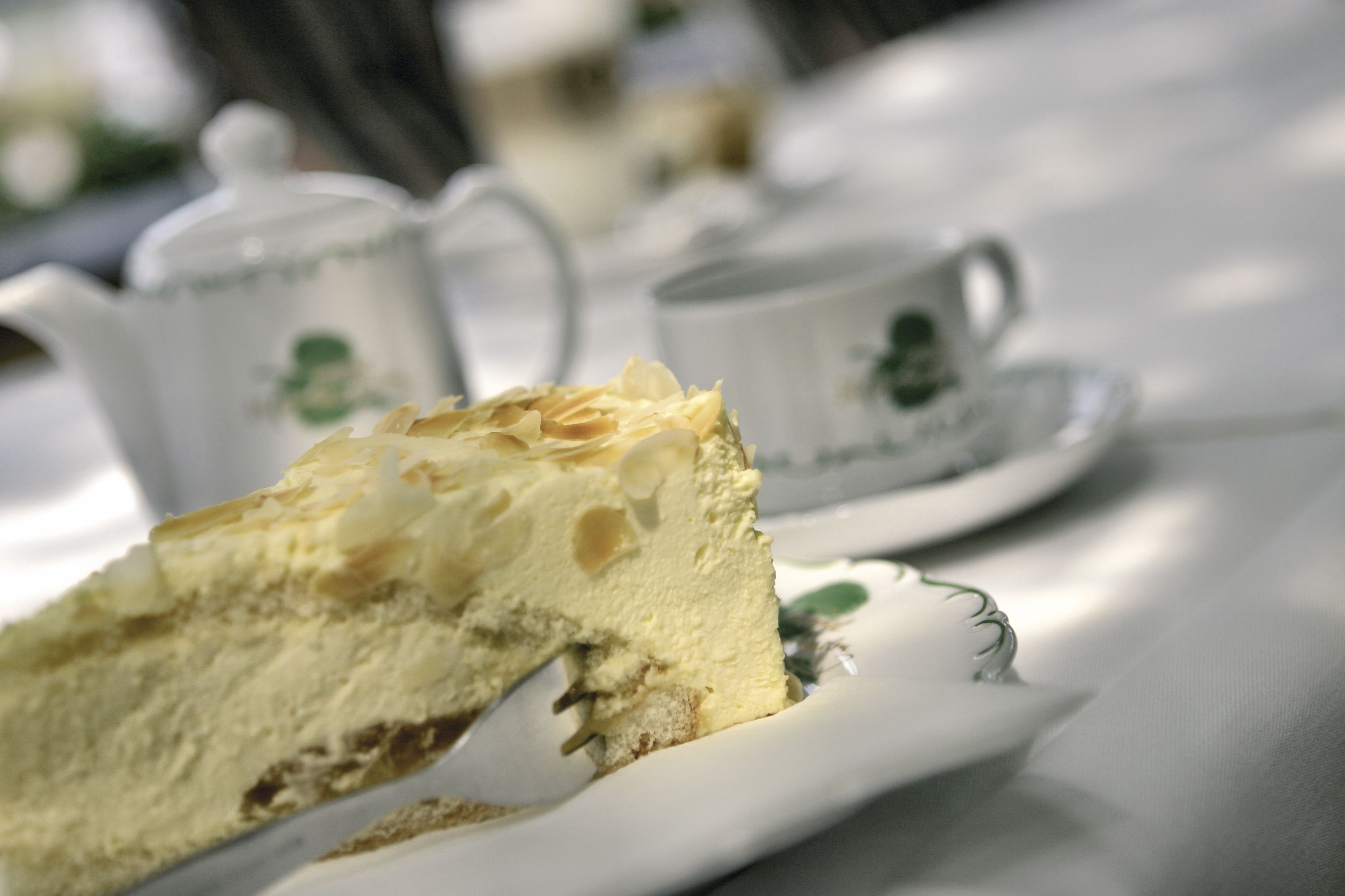 Una porción de pastel en un plato blanco con diseño verde, una tetera blanca y una taza blanca con logotipo verde sobre un mantel.