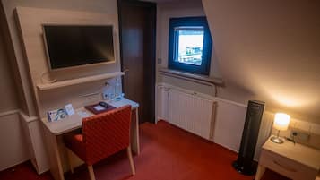 A compact room with a desk, chair, TV, window, radiator, and bedside table with a lamp.