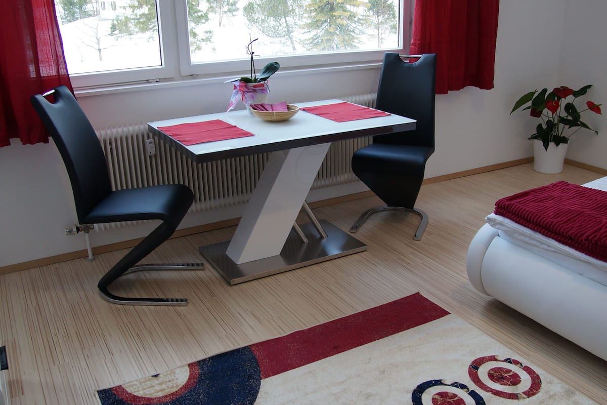 Une pièce comprenant une table, deux chaises, un radiateur, une fenêtre avec des rideaux rouges, une plante en pot et un lit recouvert d’une couverture rouge.