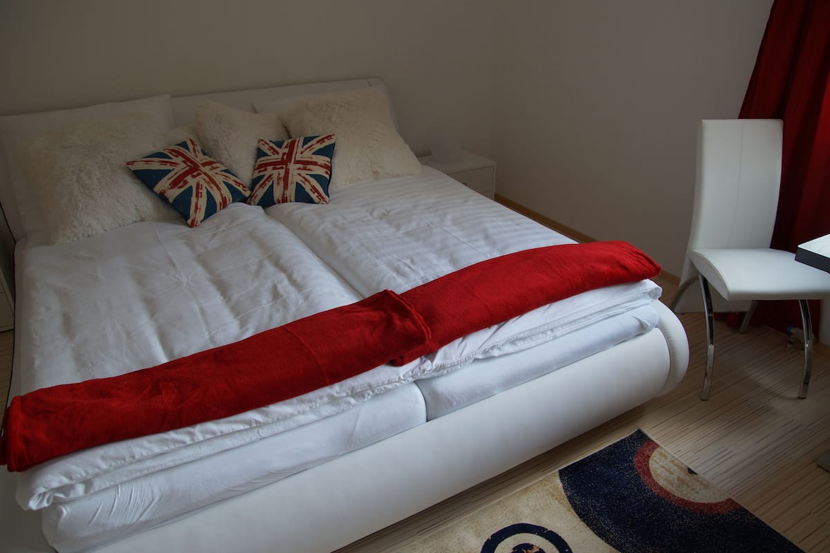 Un lit bien fait, avec du linge de lit blanc et une couverture rouge, deux oreillers ornés du motif Union Jack, une chaise blanche et une petite table de chevet.