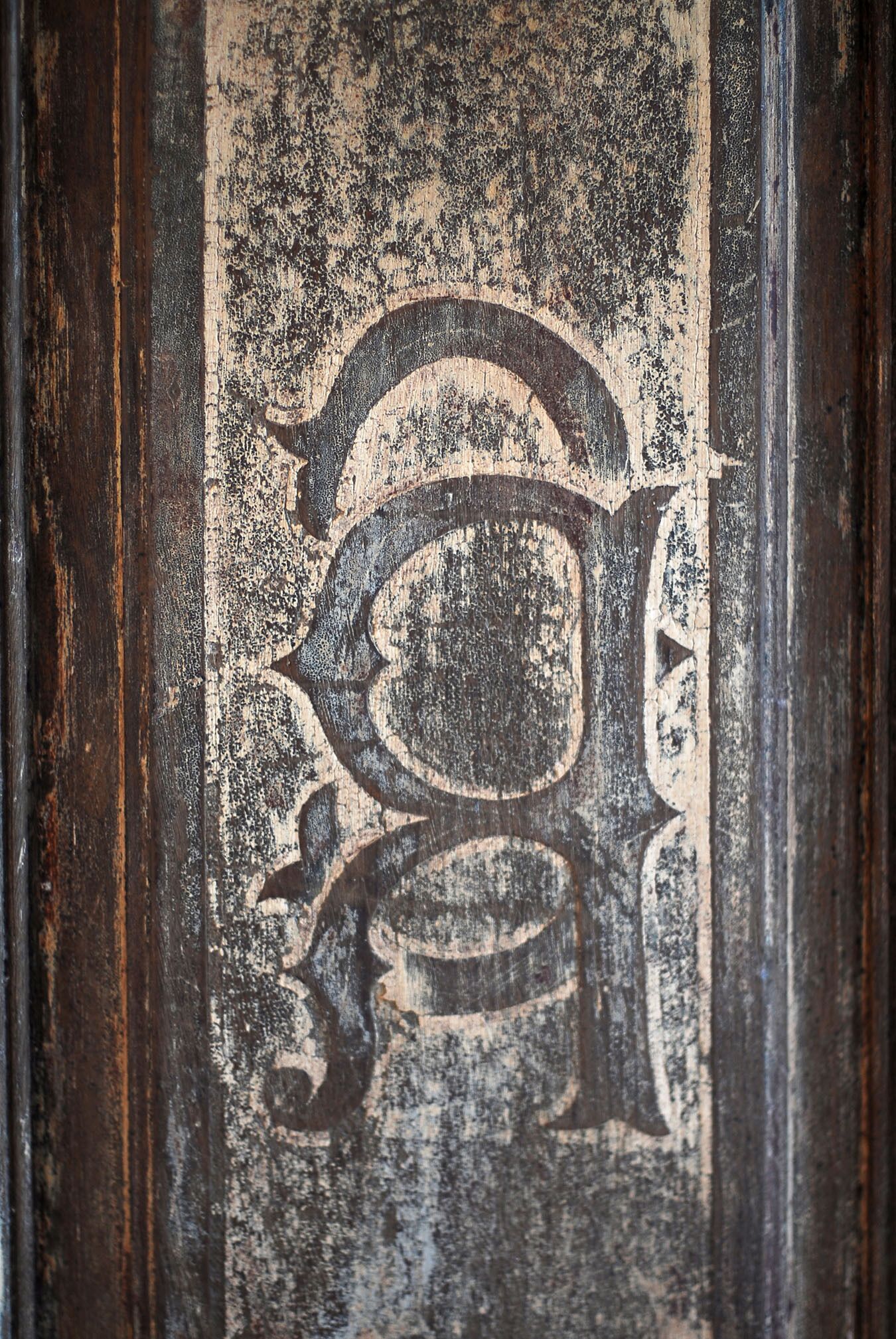 Un primo piano di una superficie di legno consunta dal tempo, con una lettera 'G' elaborata e sbiadita.