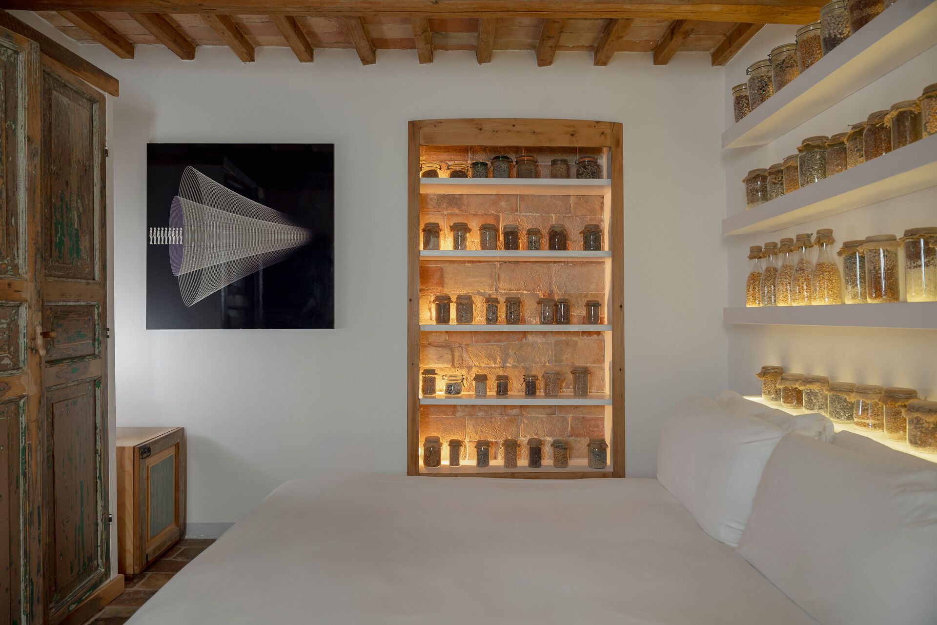 A bedroom with a bed, a wooden shelf unit with jars, a wall-mounted artwork, and a rustic door.