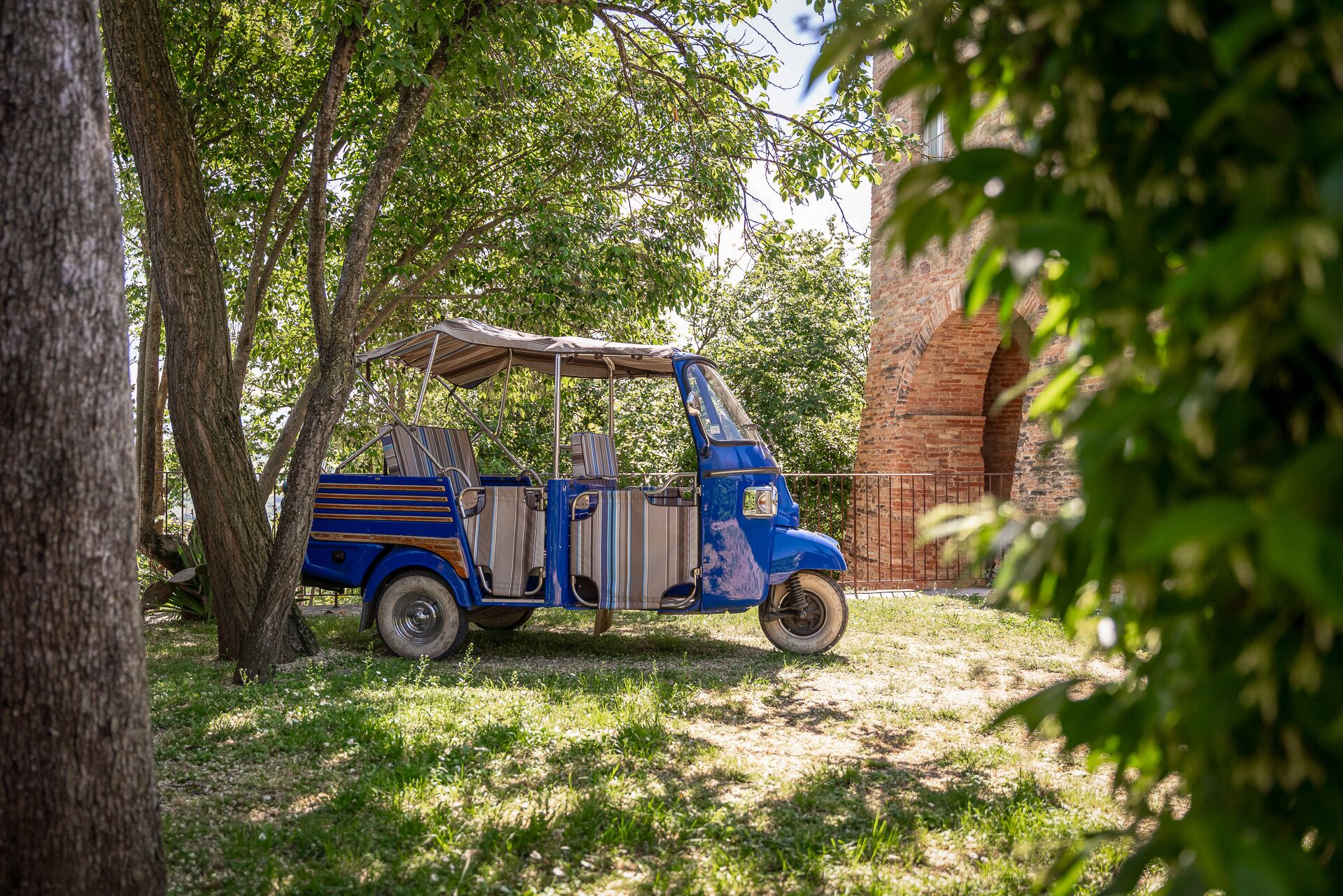 Ein blaues dreirädriges Fahrzeug steht in schattigen Baum- und Gebäudeumgebung geparkt.