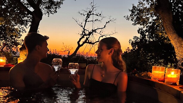 A couple in a hot tub with candles and a sunset in the background.