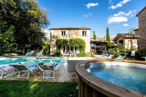 Un espace piscine doté de chaises longues et d’un jacuzzi, entouré de bâtiments en pierre et de verdure.