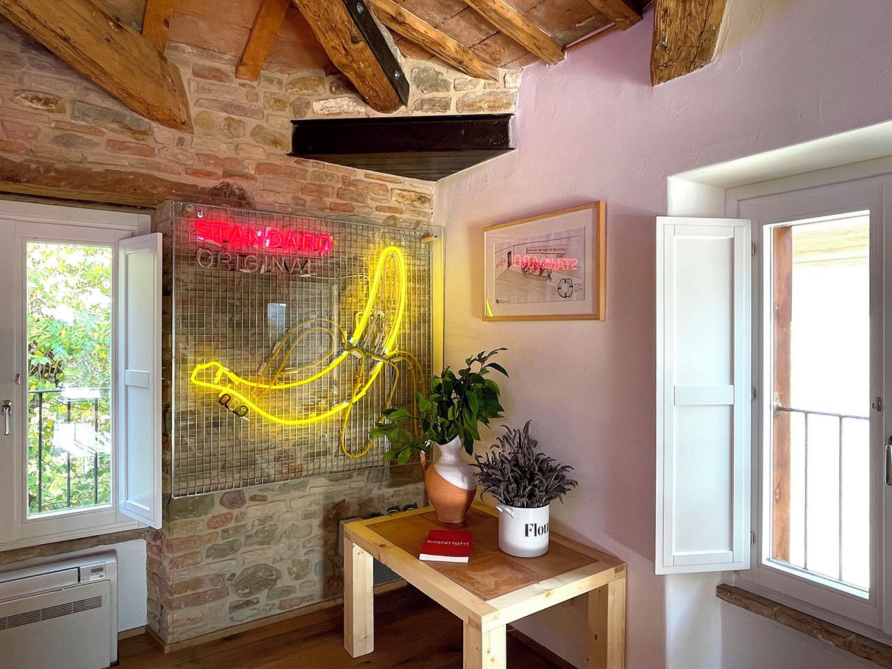 Una habitación con pared de piedra, techo de madera, una mesa con un florero y una planta, un letrero de neón y una ventana con vista a follaje.
