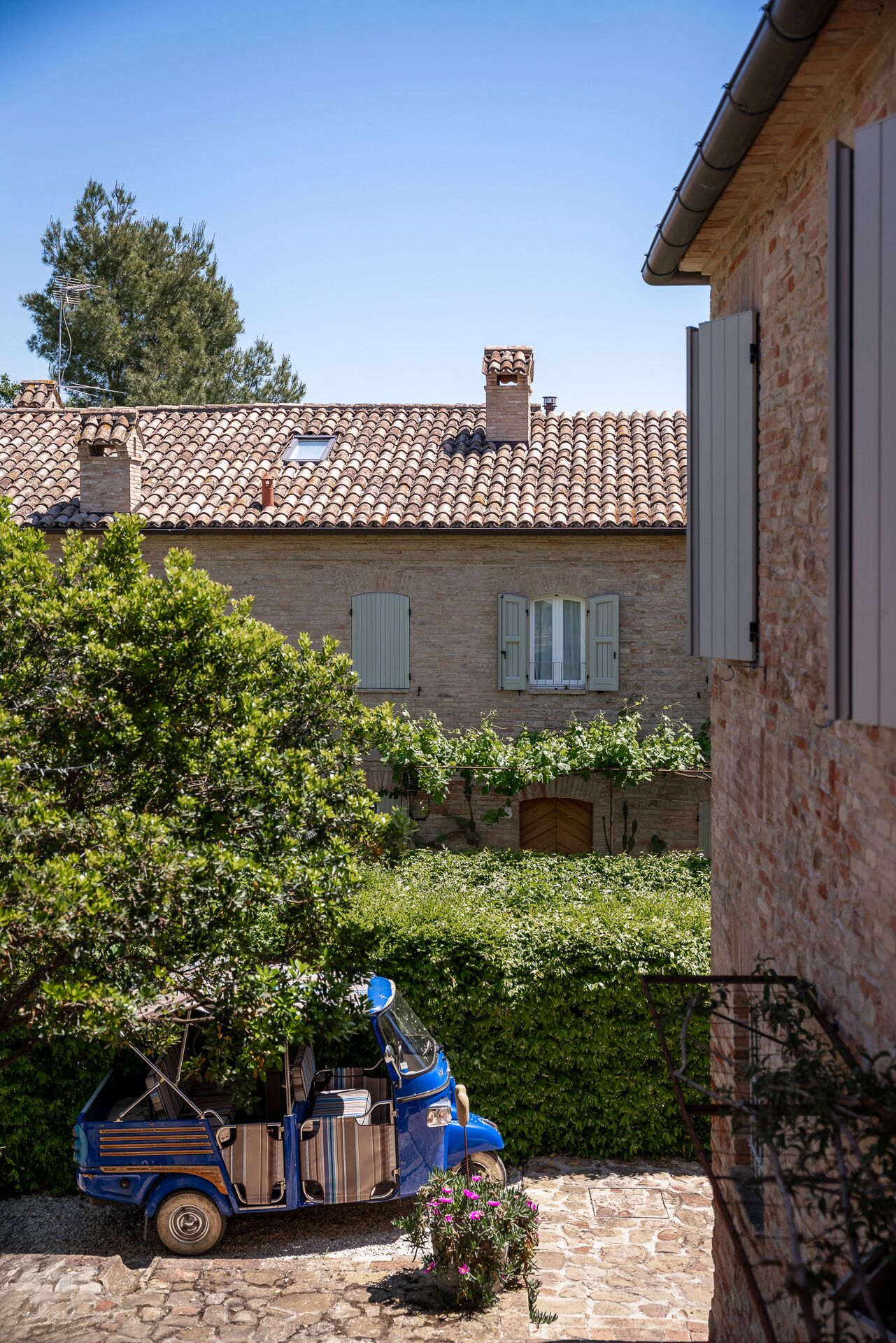 A rustic building with a tiled roof, a wooden door, and a small garden area with a blue vehicle parked in front.