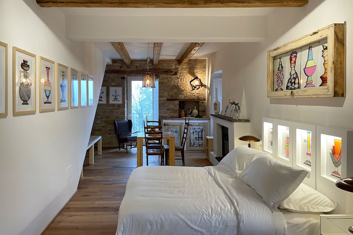 Ein Schlafzimmer mit einem großen Bett, einem Schreibtisch, Stühlen, einem Kamin und Kunstwerken an den Wänden.