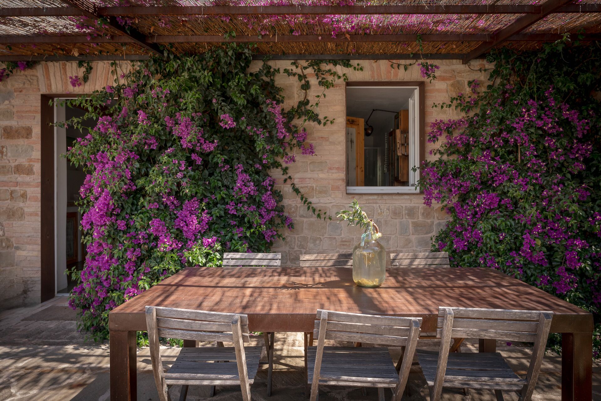 Une table en bois, des chaises, une bouteille et une fenêtre donnant sur un mur de pierre et des plantes grimpantes.