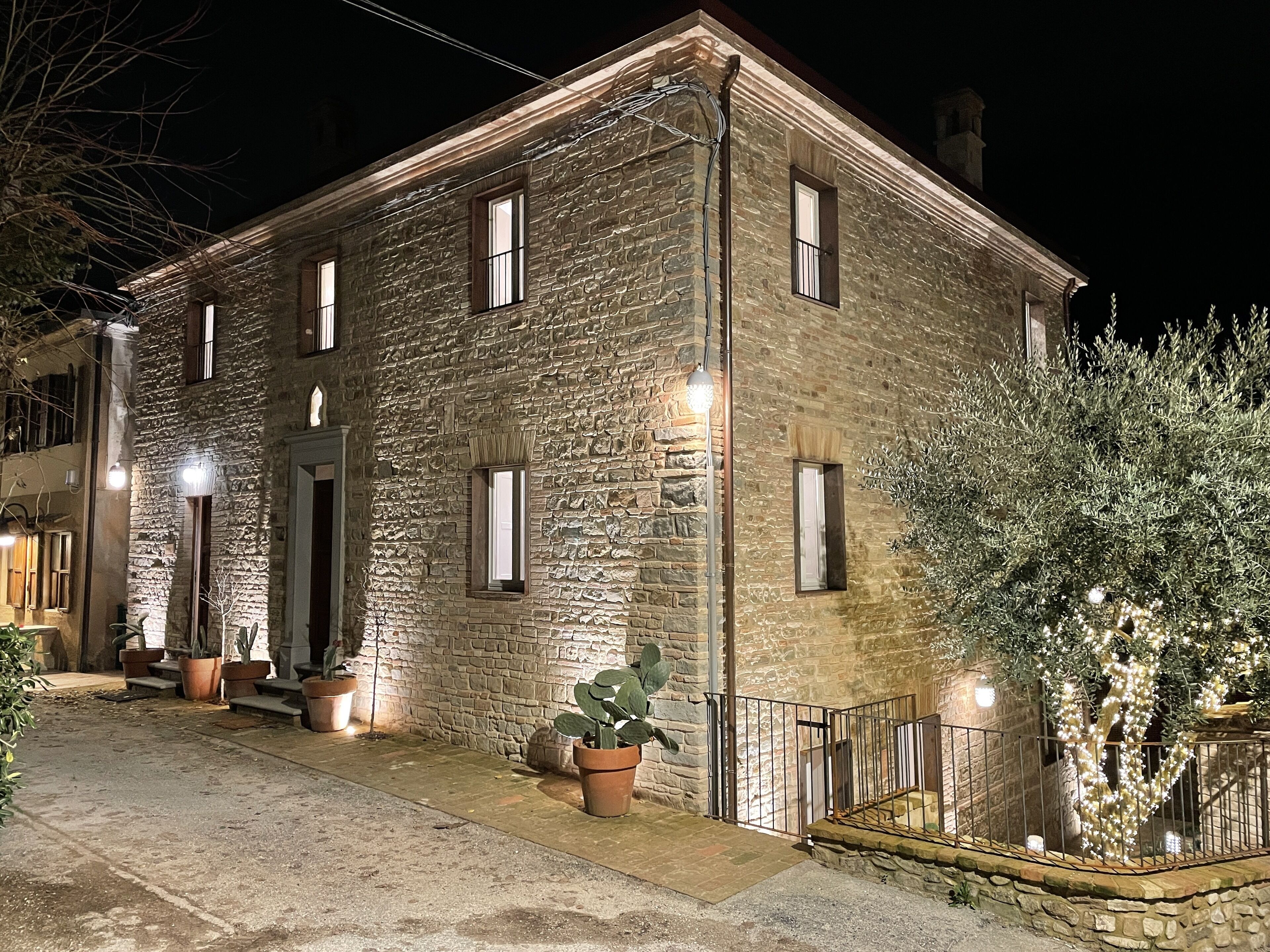 Un bâtiment en pierre doté de nombreuses fenêtres, d’une porte et d’un petit balcon avec une balustrade. Des plantes en pot et un arbre décoré de guirlandes lumineuses se trouvent sur le balcon.