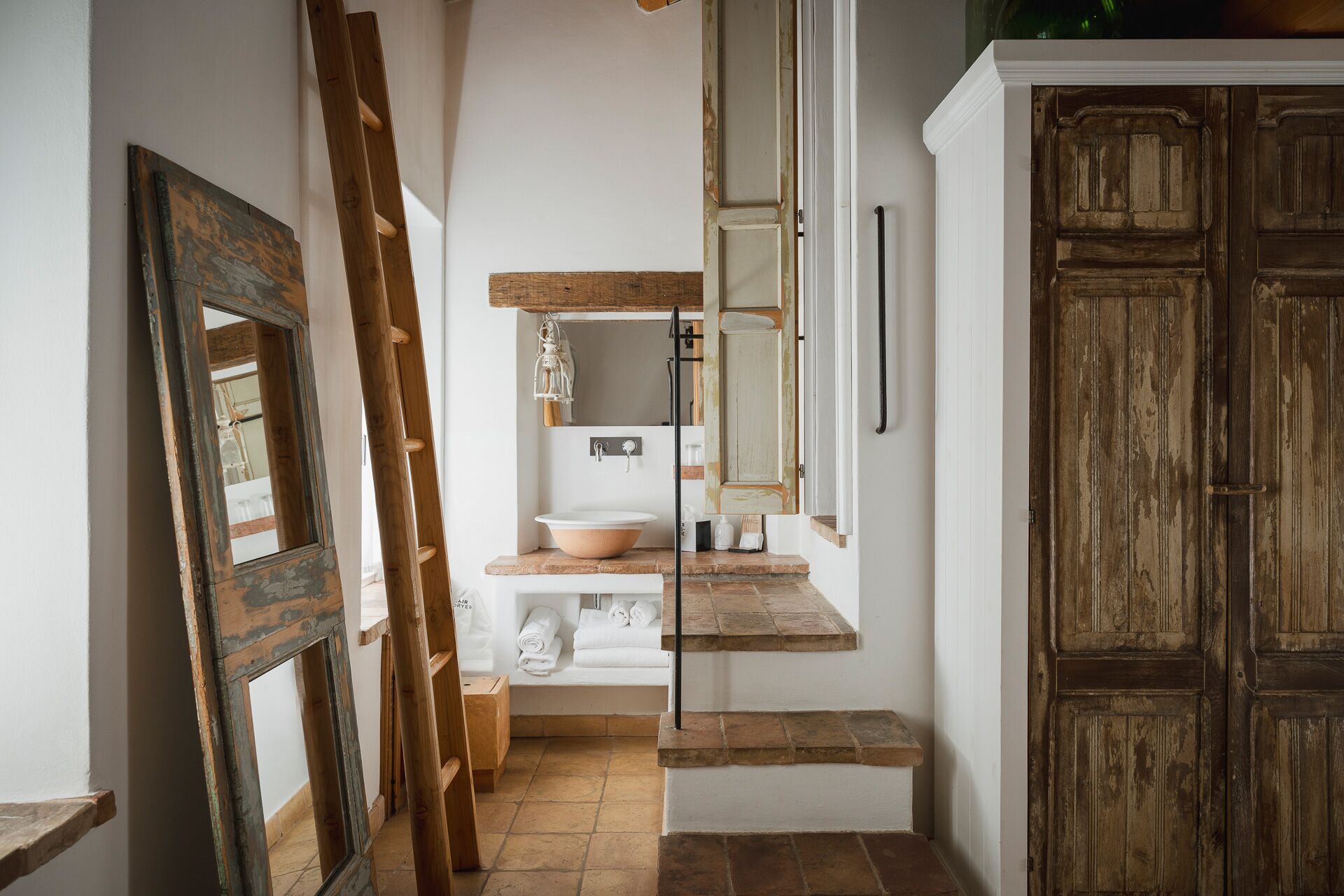 Un corridoio stretto con una porta in legno rustica, un lavandino bianco con bacino e una scala di legno appoggiata al muro.