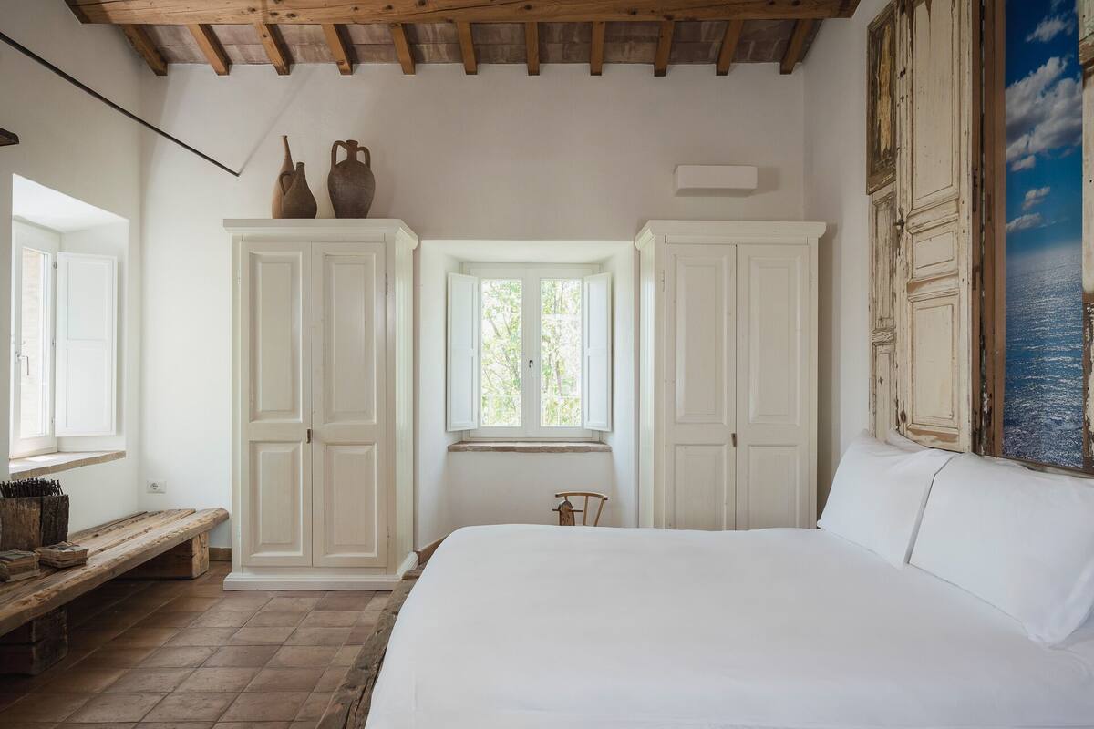 Ein Schlafzimmer mit einem großen Bett, Holzbalken an der Decke, weißen Schränken, einem Fenster und Blick zum Himmel.