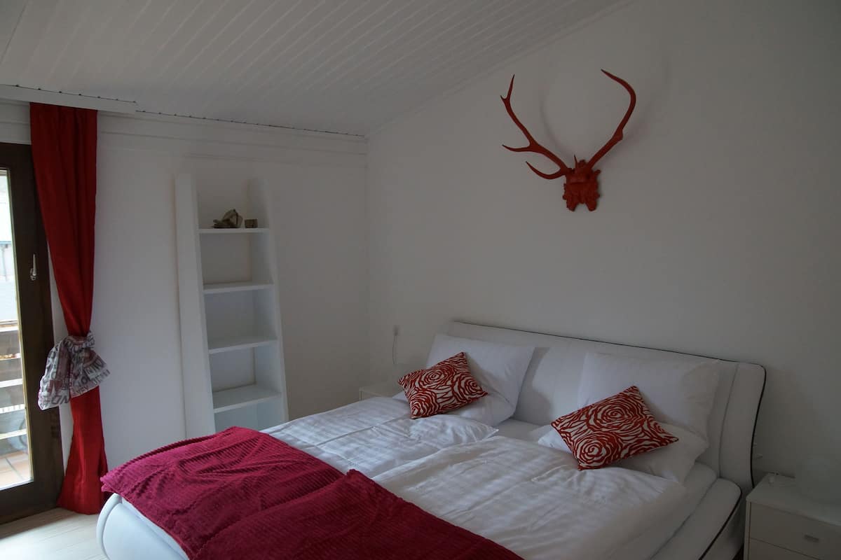 A bedroom with a bed, red and white patterned pillows, a red blanket, a white headboard, a white nightstand, and a red antler mounted on the wall.